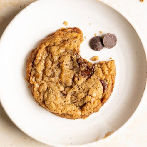 A bitten toffee sourdough chocolate chip cookie on a plate with a couple chocolate chips.