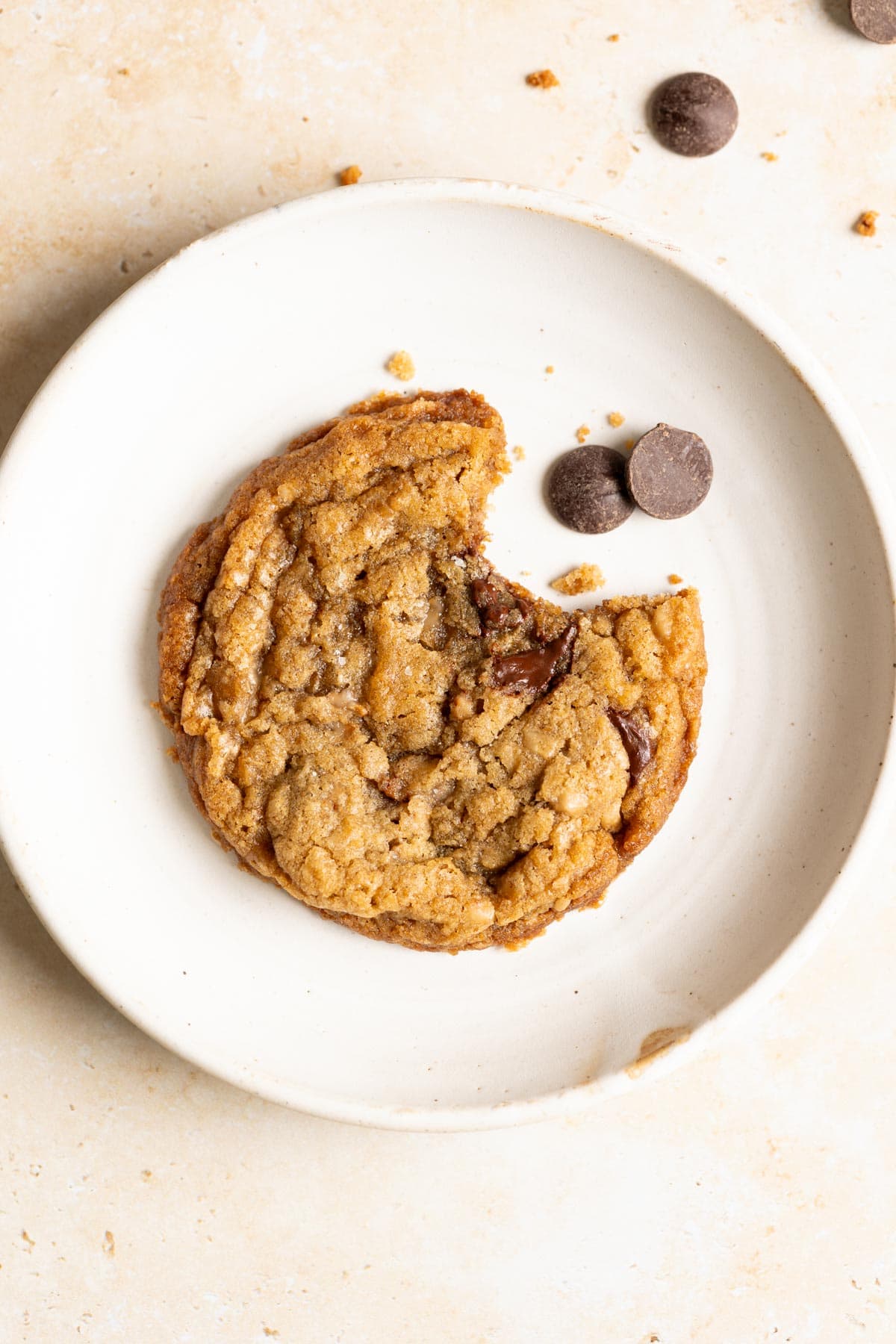 A bitten toffee sourdough chocolate chip cookie on a plate with a couple chocolate chips.