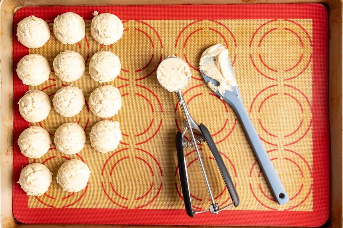 A cookie scoop with dough in it, next to a spatula, on a baking sheet with dough balls off to the side.