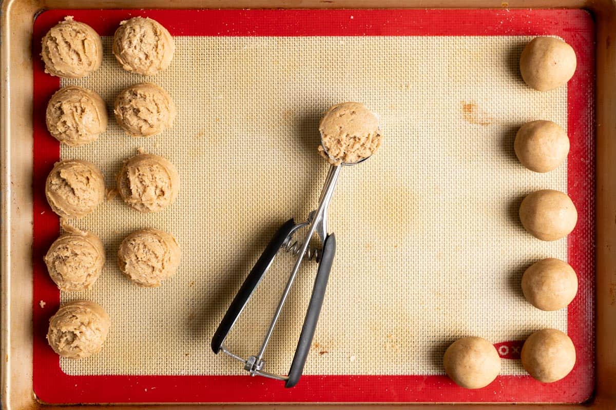 A cookie scoop with dough inside in the middle of a baking sheet with dough balls on both sides.
